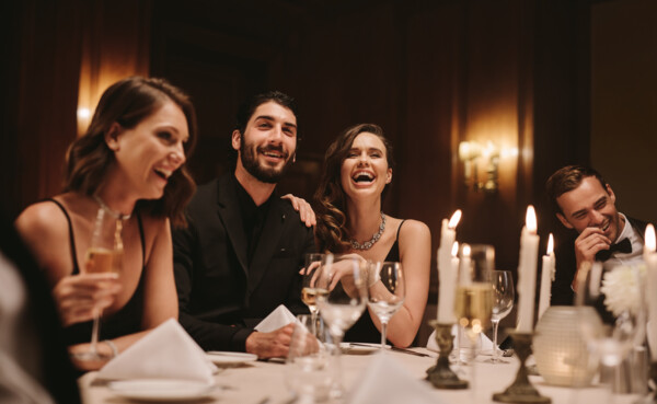 Elegante Gruppe von Menschen in Abendgarderobe sitzt an einem festlich gedeckten Tisch, lacht gemeinsam und genießt ein luxuriöses Silvester-Dinner bei Kerzenschein. © 